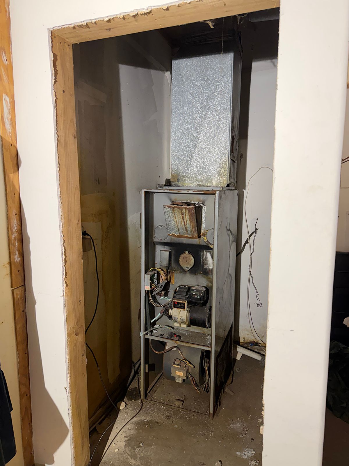An old, dismantled HVAC furnace sits inside a narrow, unfinished utility closet with exposed concrete flooring.