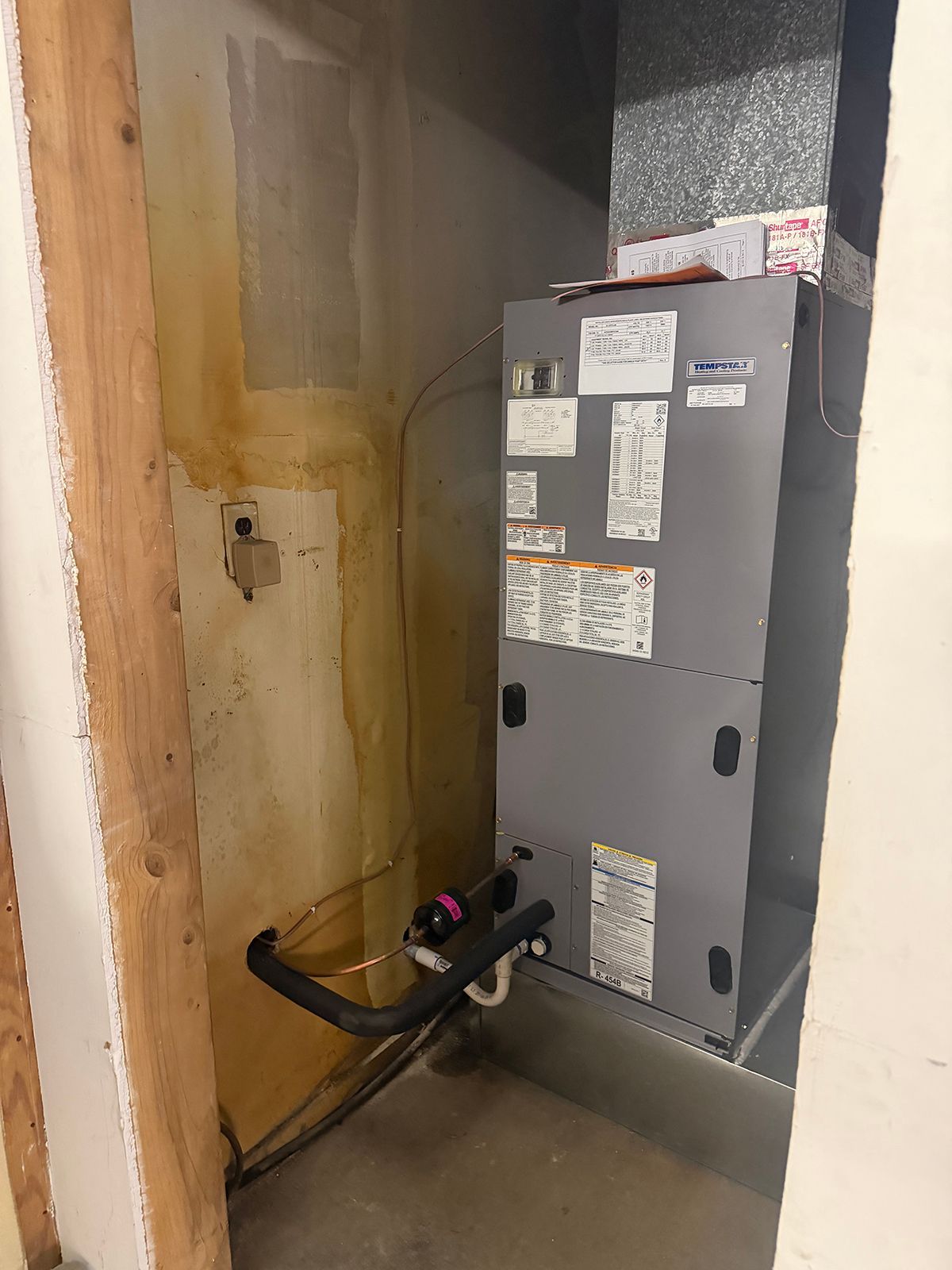 A gray HVAC air handler unit standing in a basement corner next to an unfinished wall with exposed wooden studs.