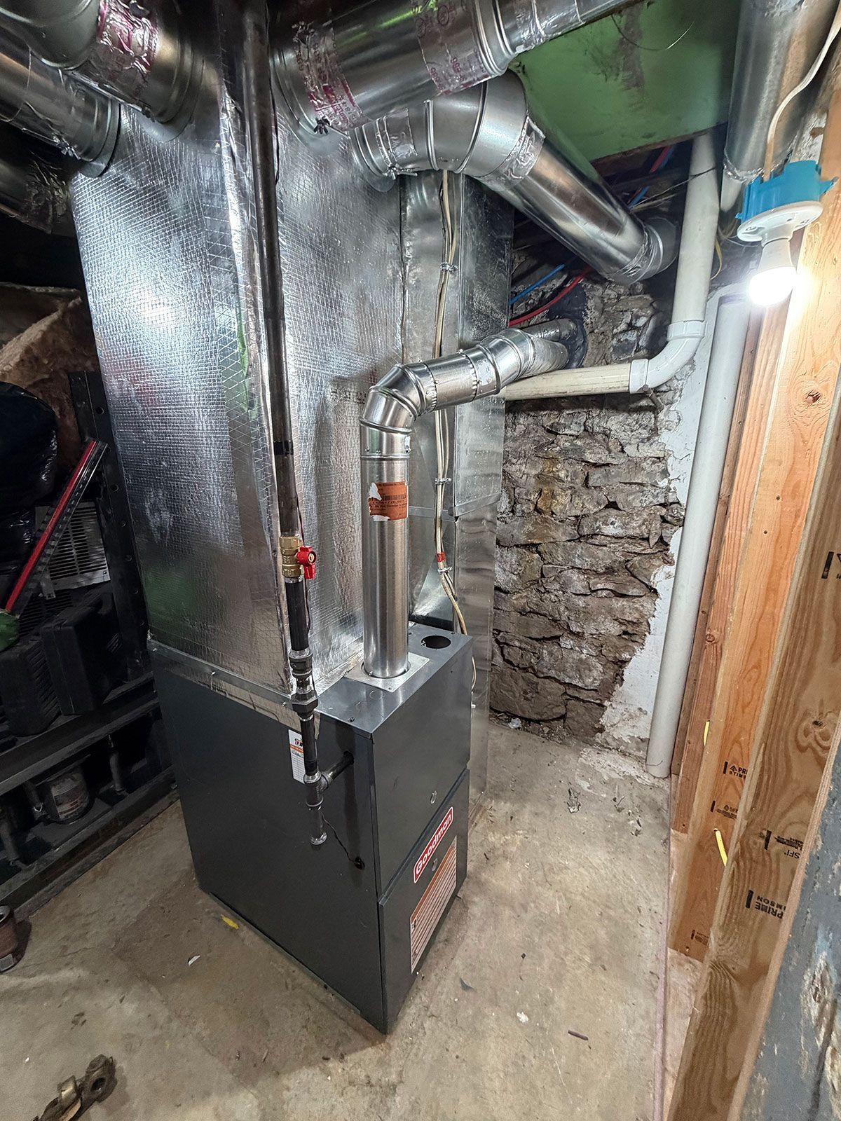 A gray furnace with silver ductwork stands in a basement against an exposed stone and white painted wall.