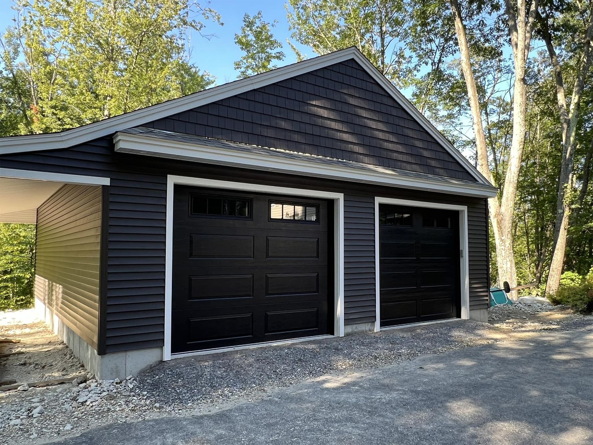 Two-car black garage with white trim. Dark gray siding, gravel driveway, and trees.