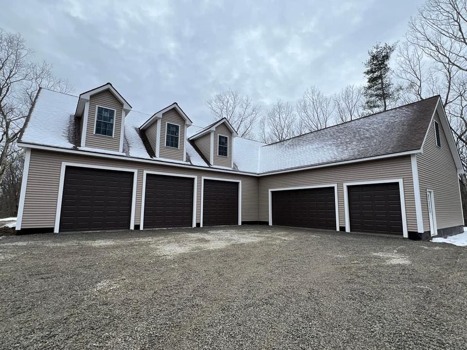 A large garage with six dark brown doors, beige siding, and three dormers with windows; snowy setting.