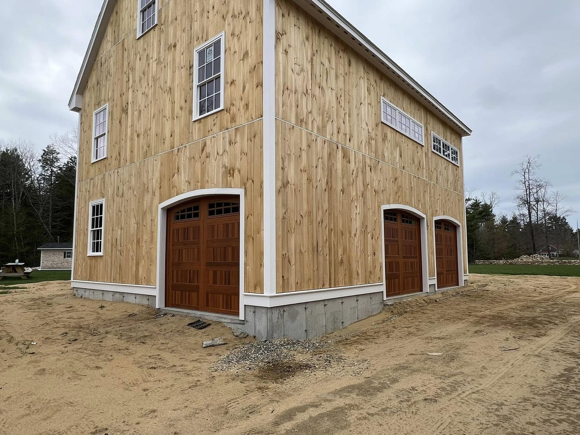 Two-story wood barn with brown garage doors and white trim, under construction in a sandy area.