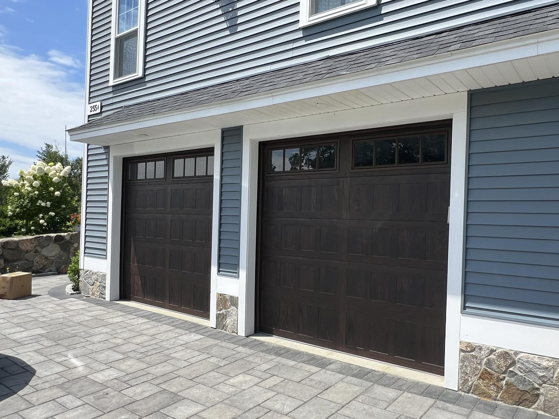Two dark brown garage doors on a blue-sided house with white trim and stone accents on a paved driveway.