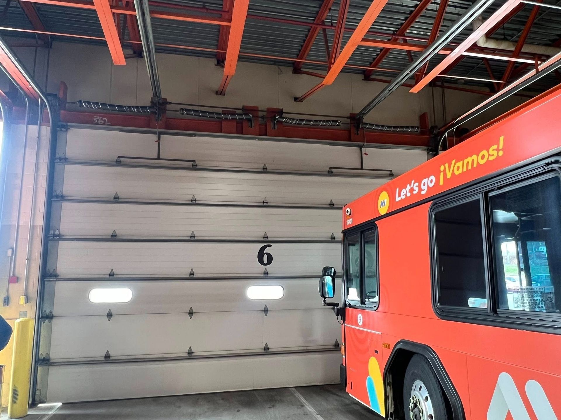 A red bus in bay number 6 at a bus depot, with a partially open garage door and orange support beams.