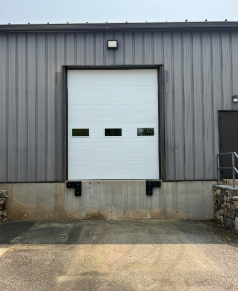 A white loading dock door on a gray industrial building with black bumper stops; asphalt in foreground.