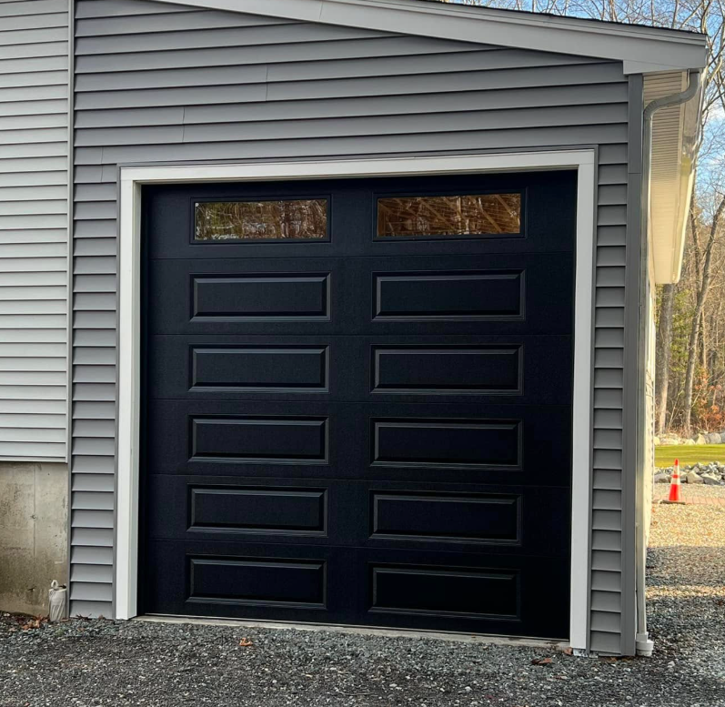 Black garage door with rectangular windows and white trim on a gray building.