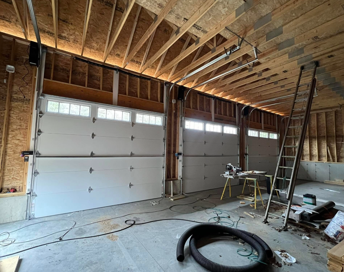 Three white garage doors in a new construction garage with exposed wooden beams and a ladder.