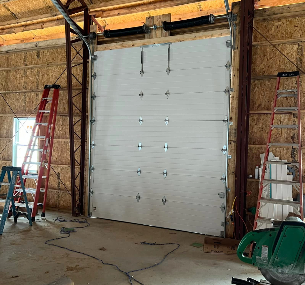 White garage door installed in a wood-framed building under construction; two red ladders flank the door.