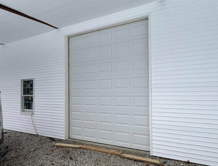 White garage door on a building with white siding and a small window. Outdoors with gravel ground.