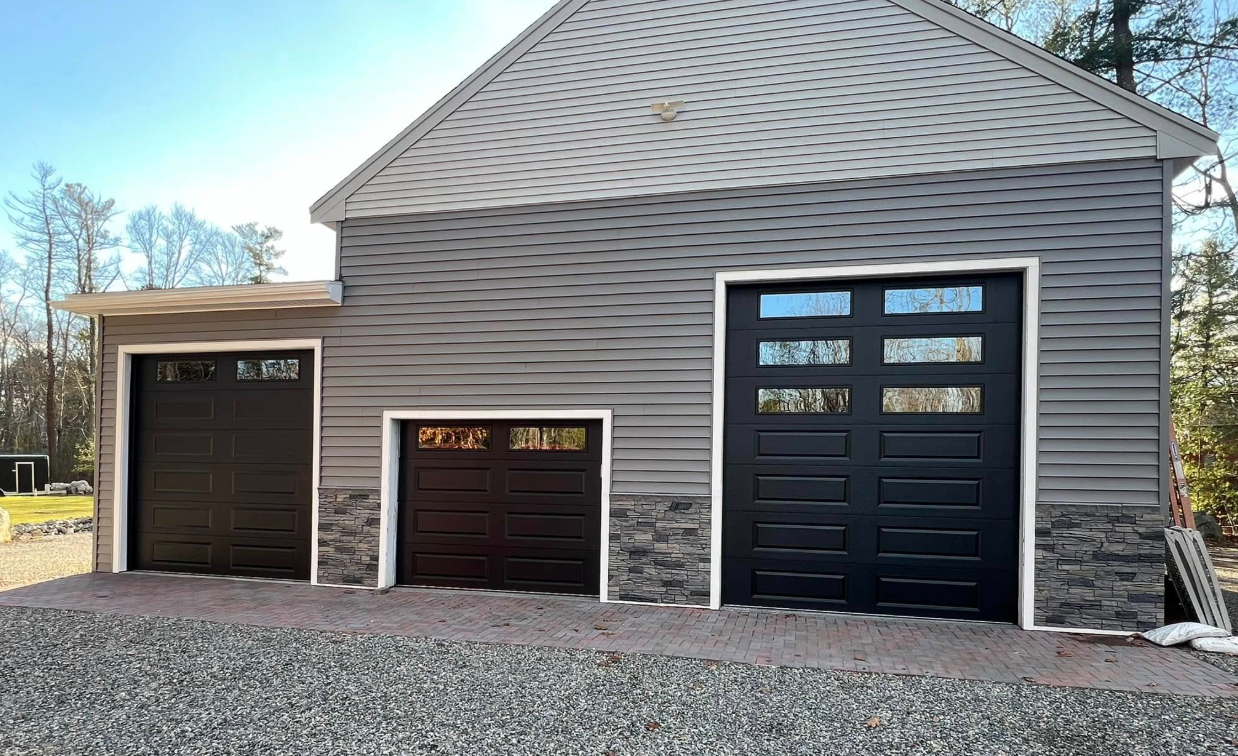 Three dark garage doors of different sizes, on a gray building, built on gravel.