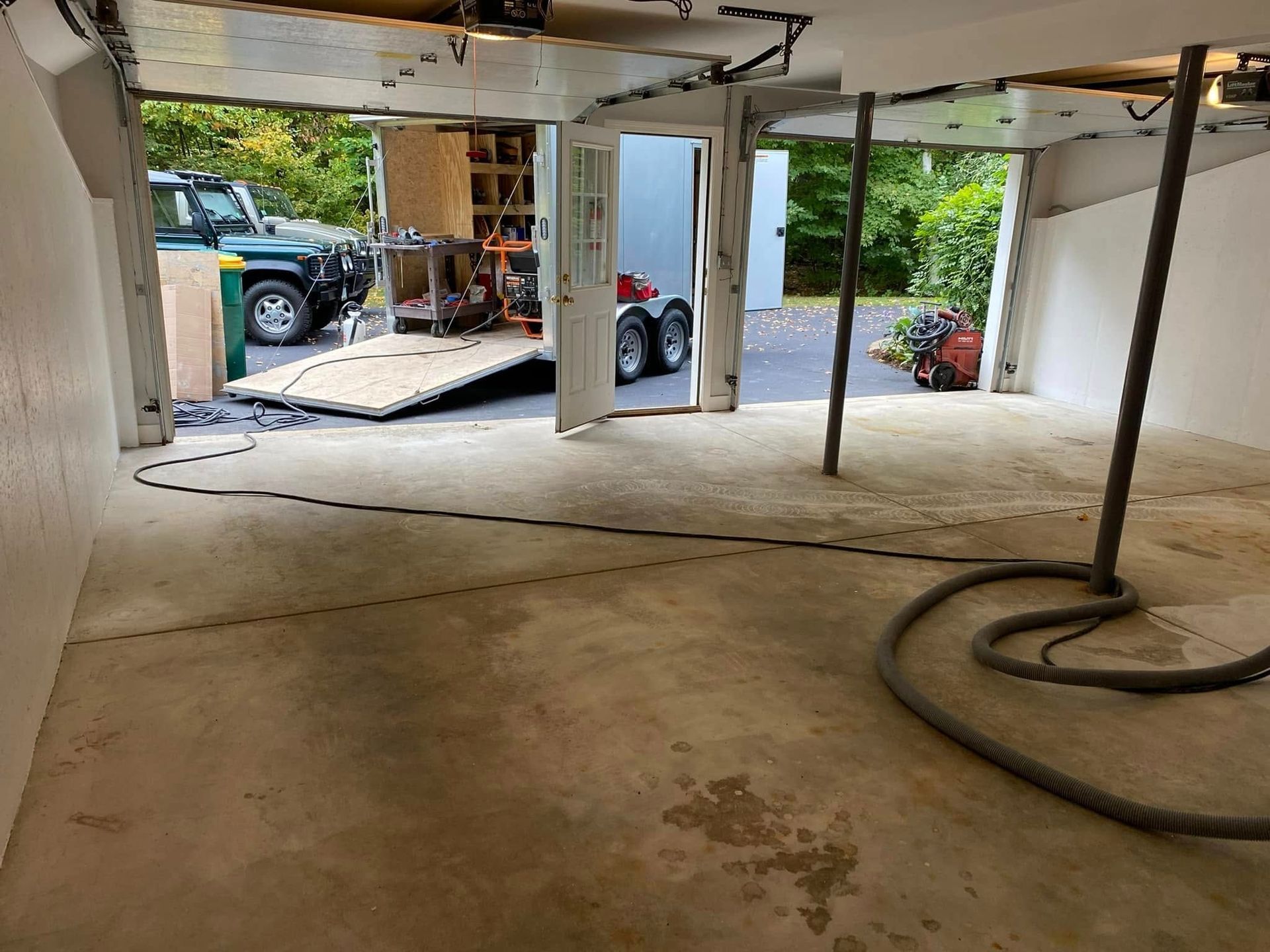 Garage interior with equipment, truck, and trailer parked outside. Concrete floor, white walls, and support beams.