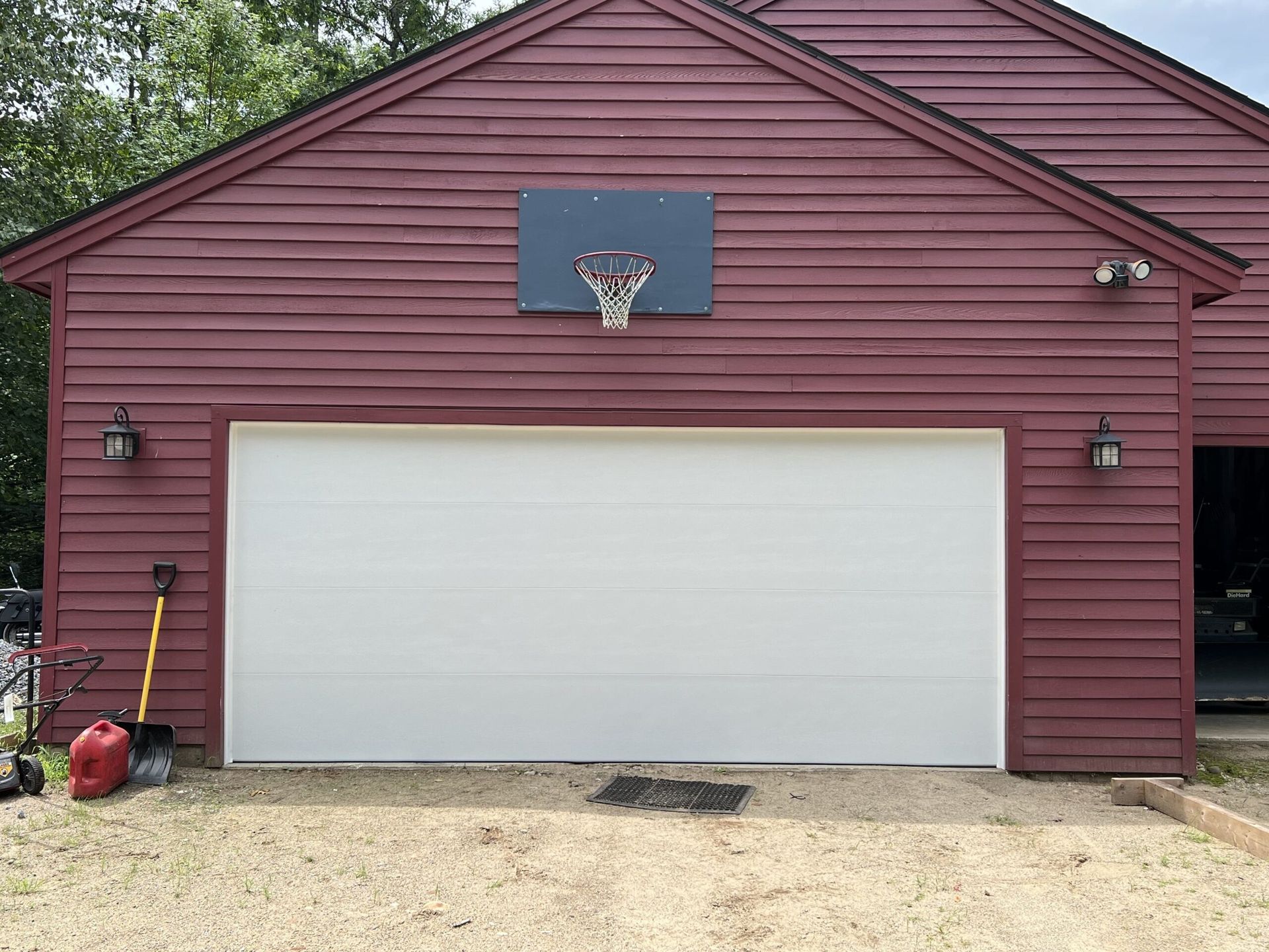 Red garage with white door, basketball hoop above.  Tools and gas can on the gravel driveway.