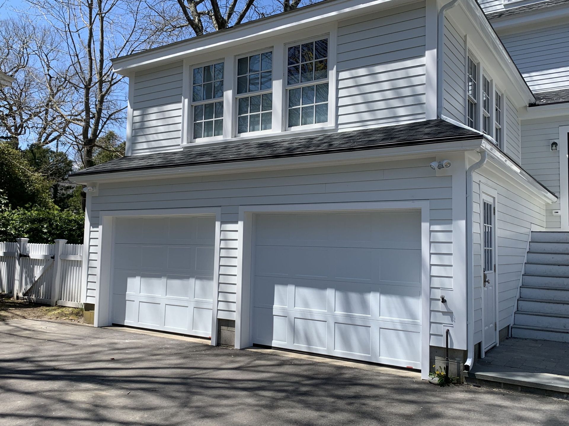 White two-car garage with upper level windows and a side entrance, located next to a white picket fence.