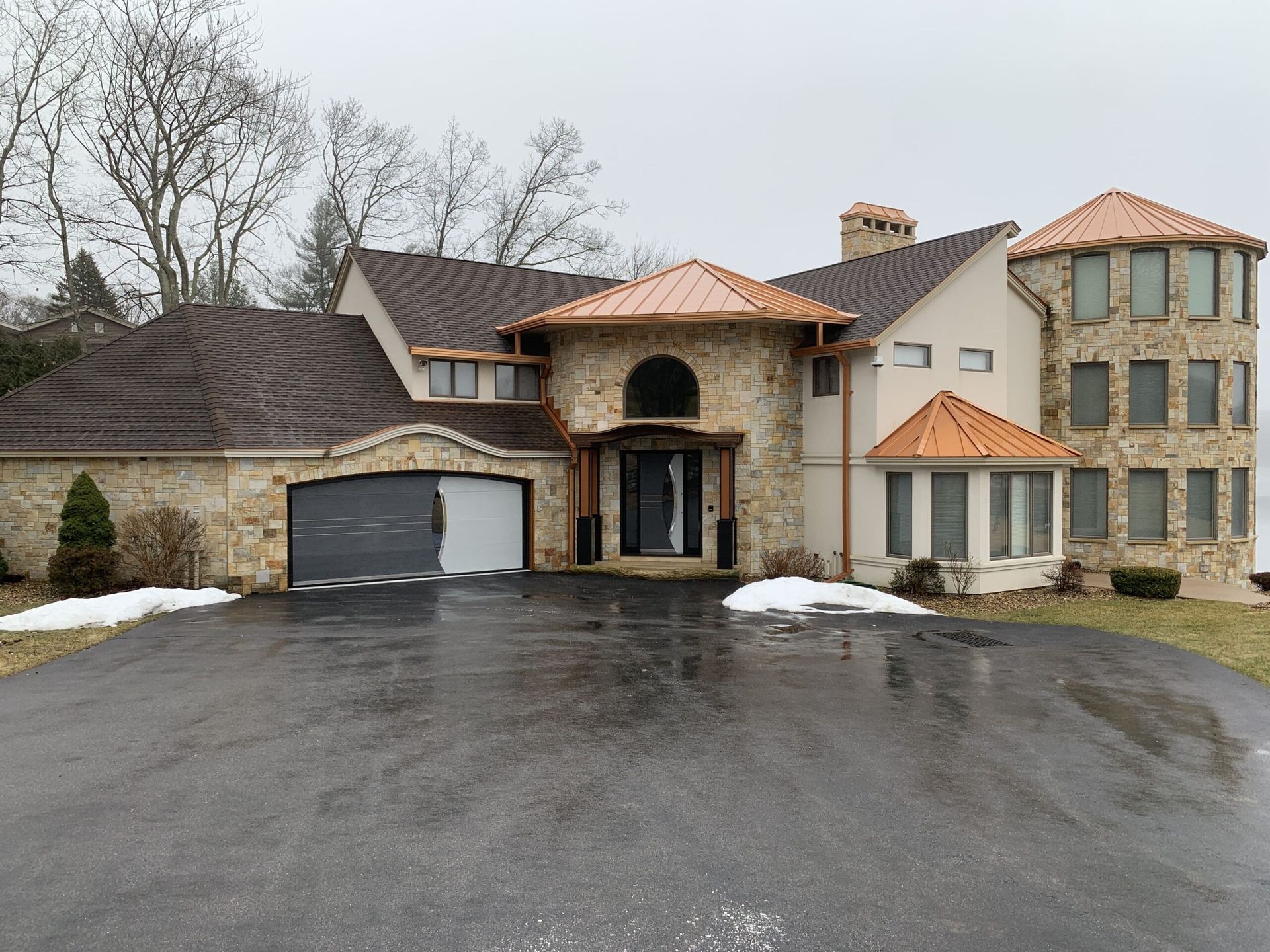 Two-story stone house with a dark driveway, brown roof, and a touch of snow.