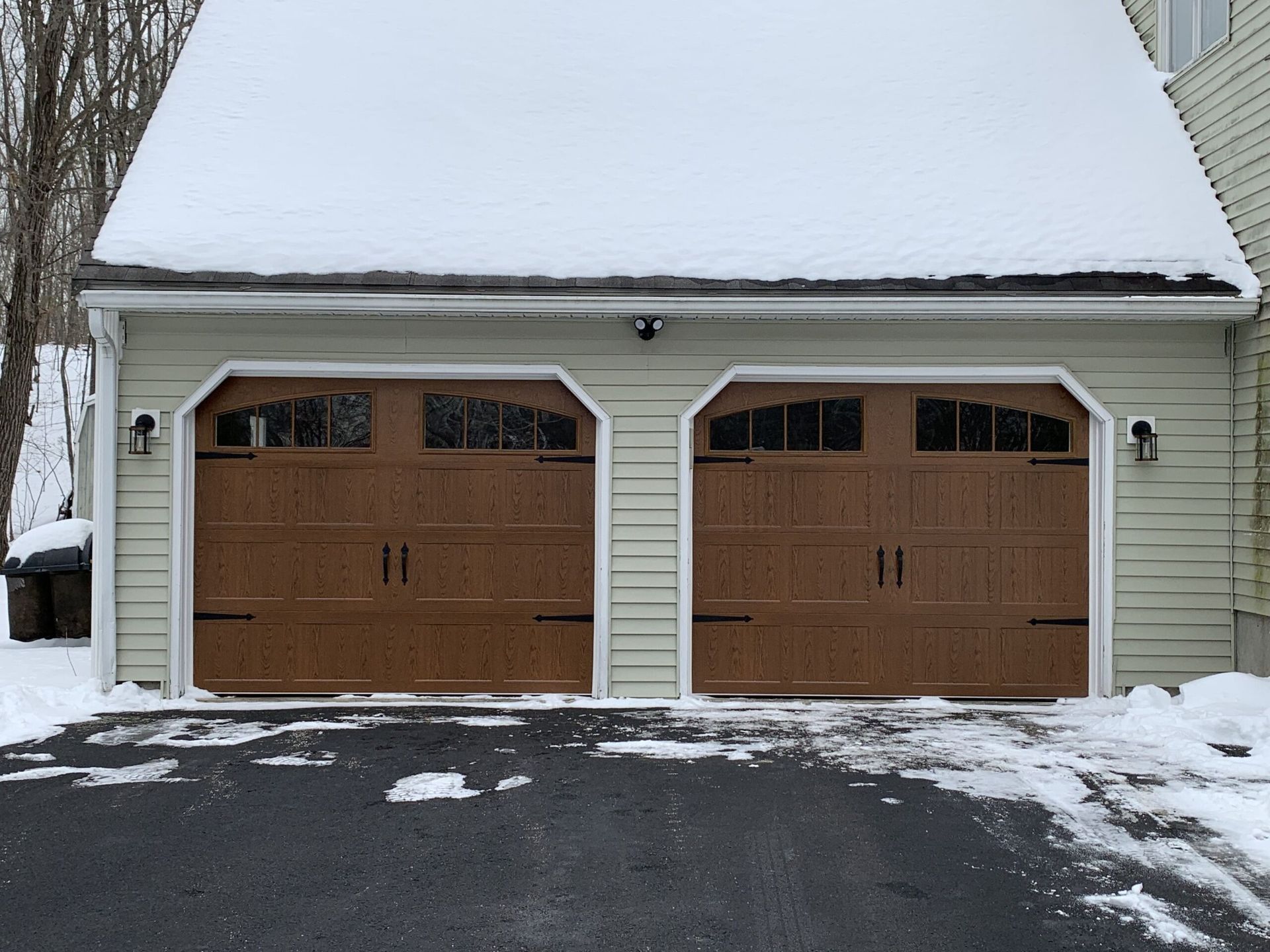 Two brown garage doors with decorative windows and hardware, snow on roof and driveway.