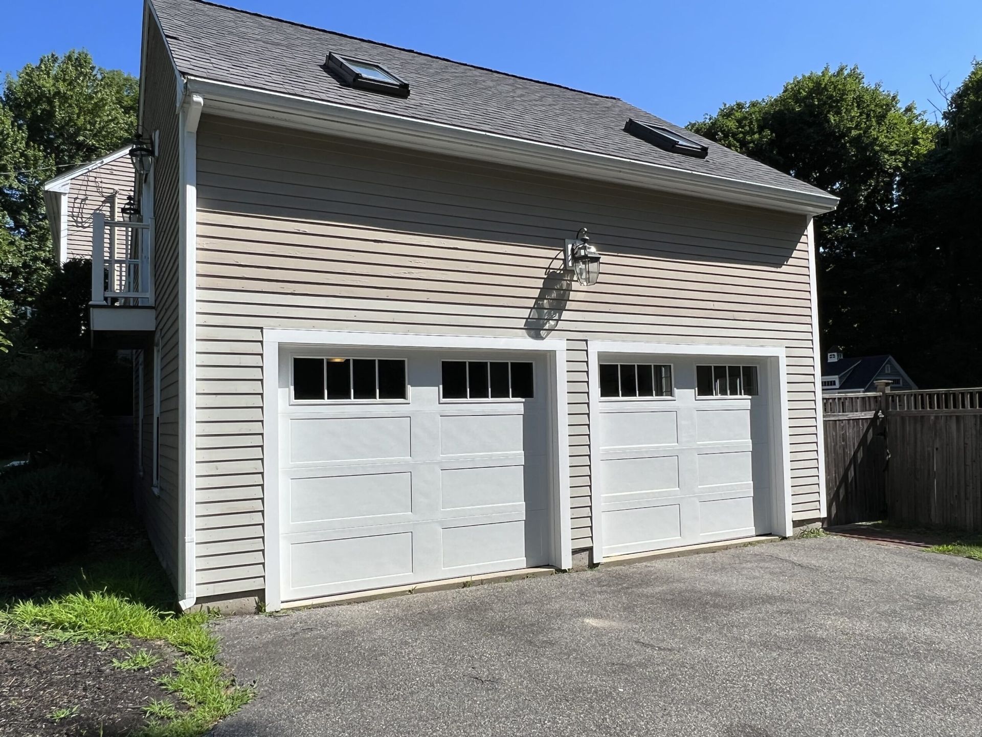 Two-car garage with white doors, light brown siding, and a balcony. Paved driveway and blue sky.
