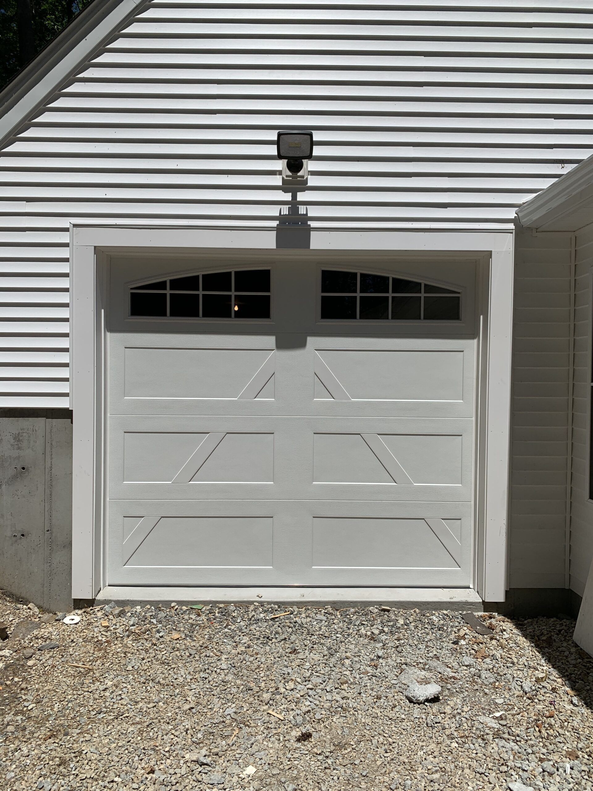 White garage door with decorative paneled design, mounted on a building with white siding and gravel ground.
