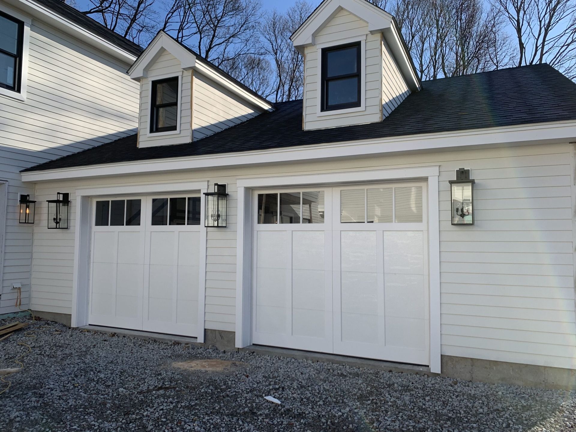 White two-car garage with black accents. Dormers, lanterns, and gravel driveway.