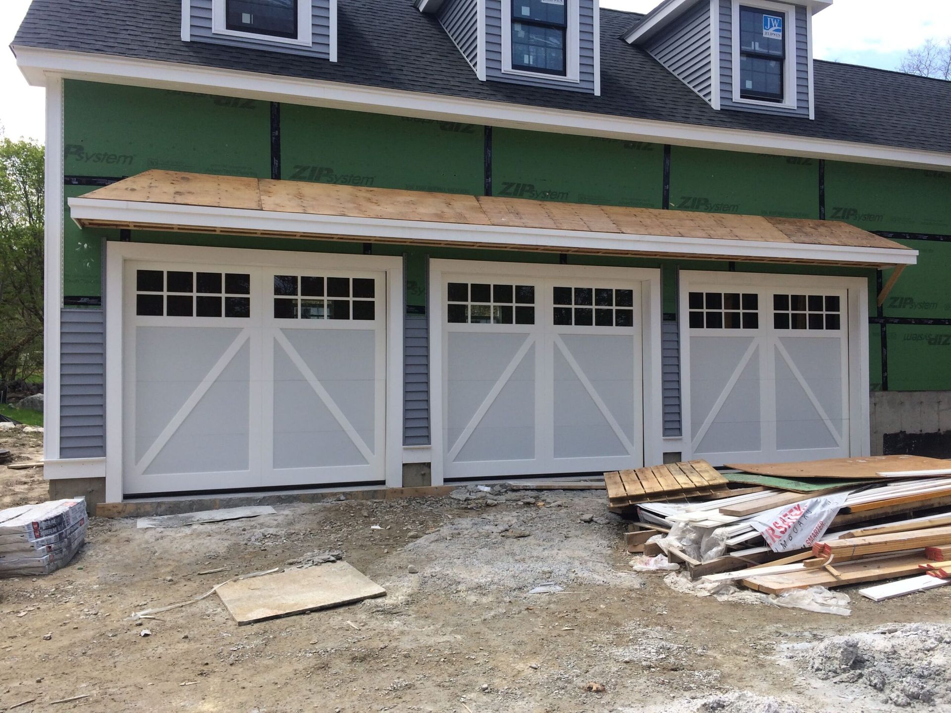 Three white garage doors with windows, under a wooden awning, at a house under construction.