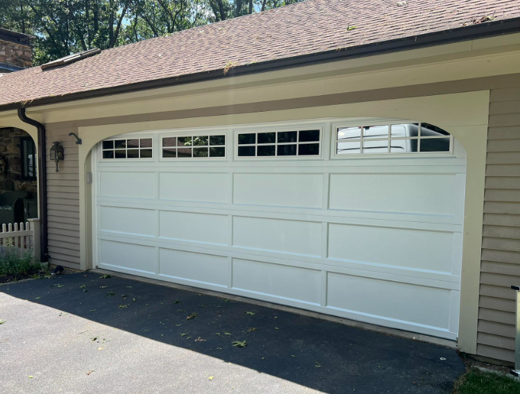 White garage door with windows on a beige house. Driveway in front, brown roof overhead.