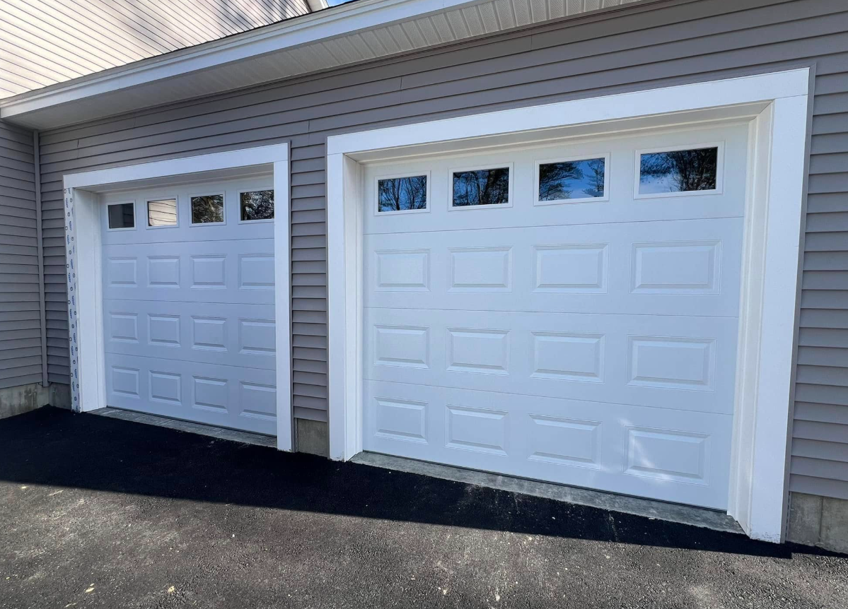 Two white garage doors with windows, bordered by white trim, on a gray-sided house. Asphalt driveway.