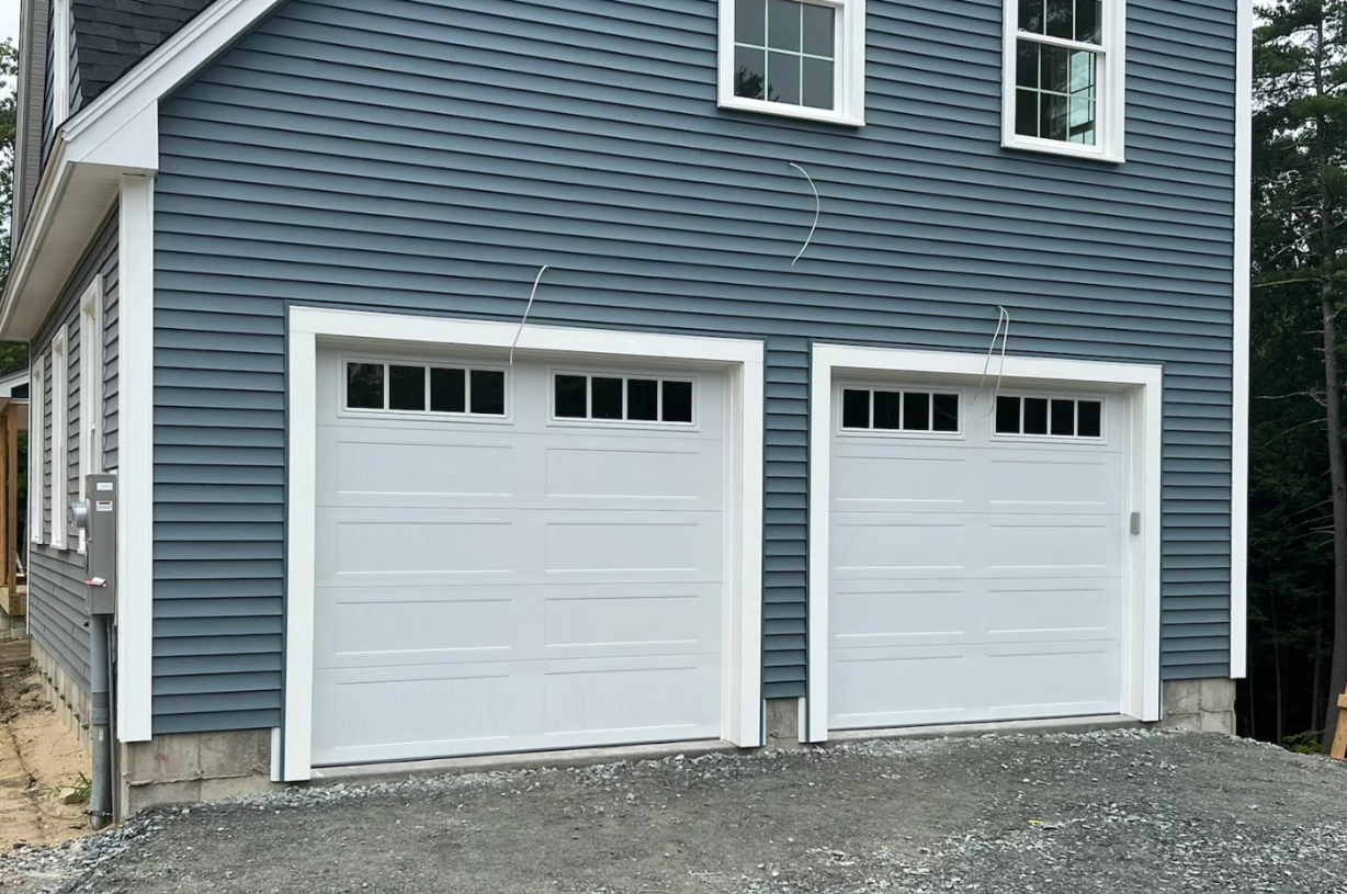 Two white garage doors with windows, against a blue-gray shingled house.