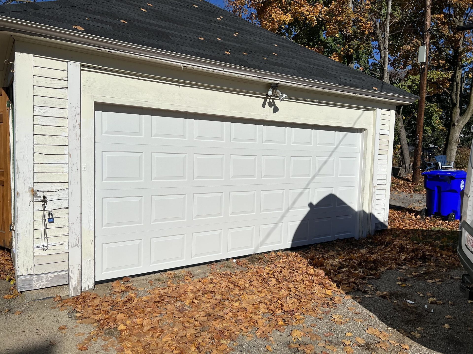 White garage door with fallen leaves in front, a blue trash bin, and some trees.