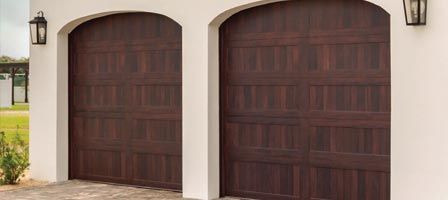 Two dark brown wooden garage doors with arched white trim and black lanterns.