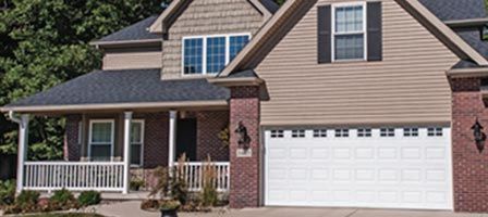 A two-story house with a white garage door, tan siding, brick accents, and a covered porch.