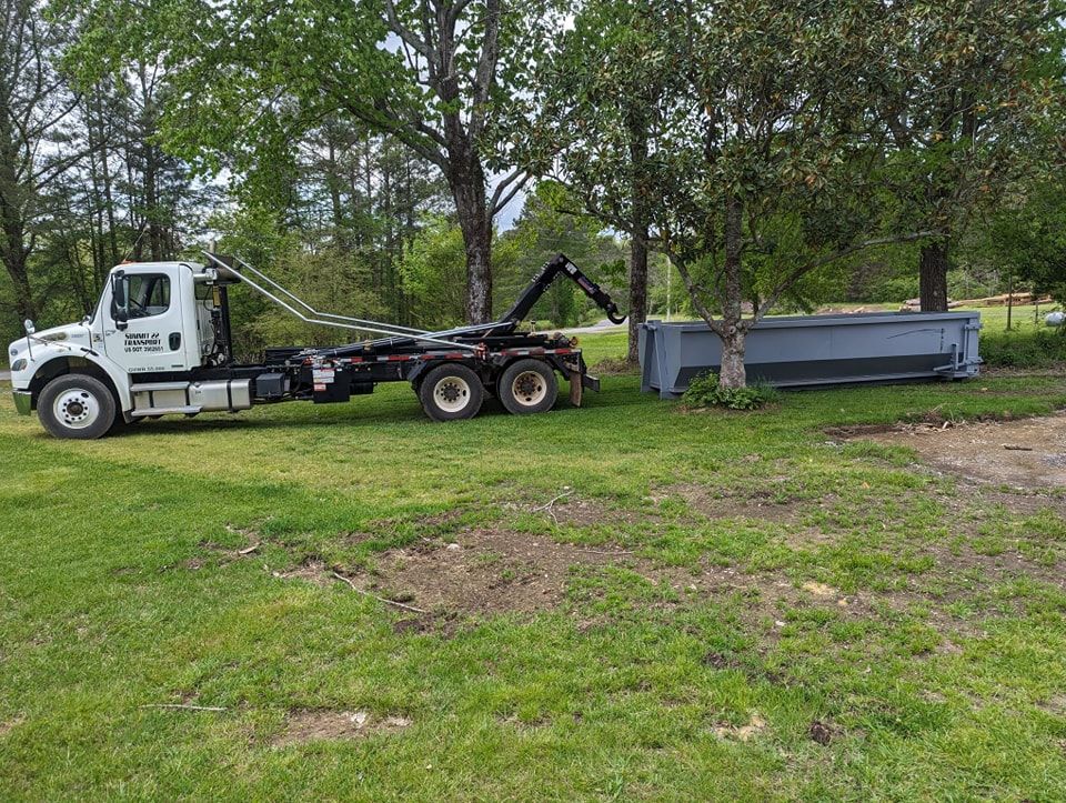 A dumpster is being towed by a dump truck in a grassy field.