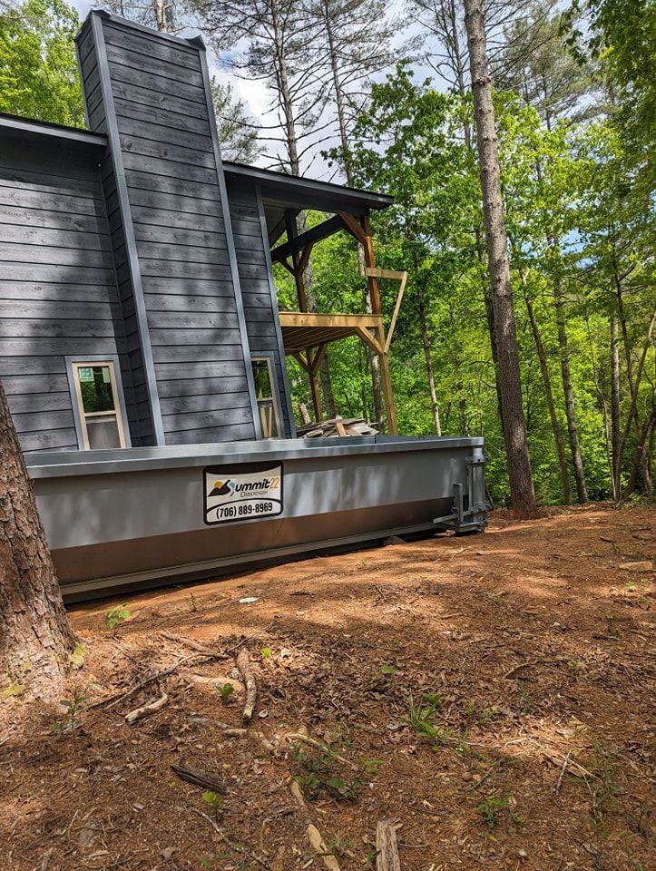 A dumpster is parked in front of a house in the woods.