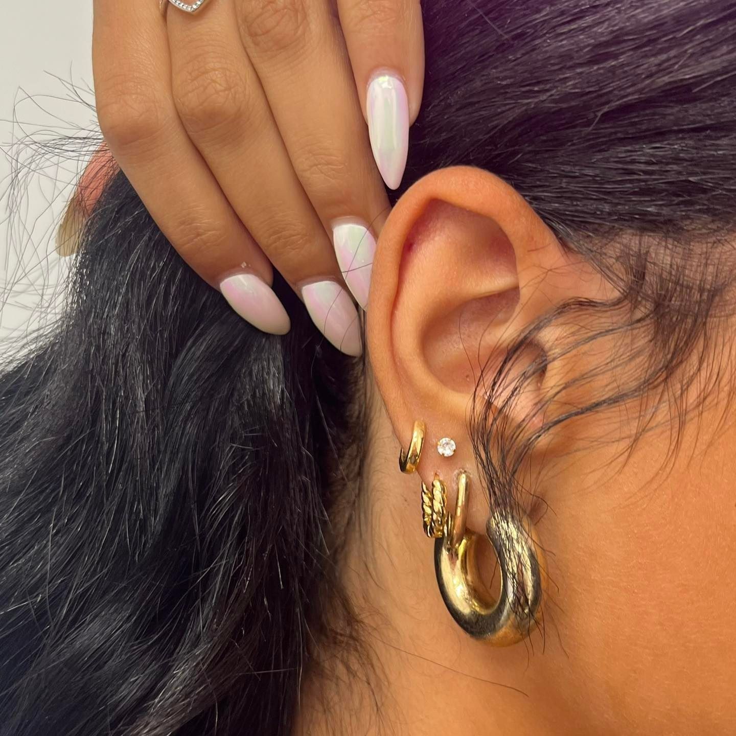 Ear with multiple gold earrings; hand with long, light pink nails resting on head.