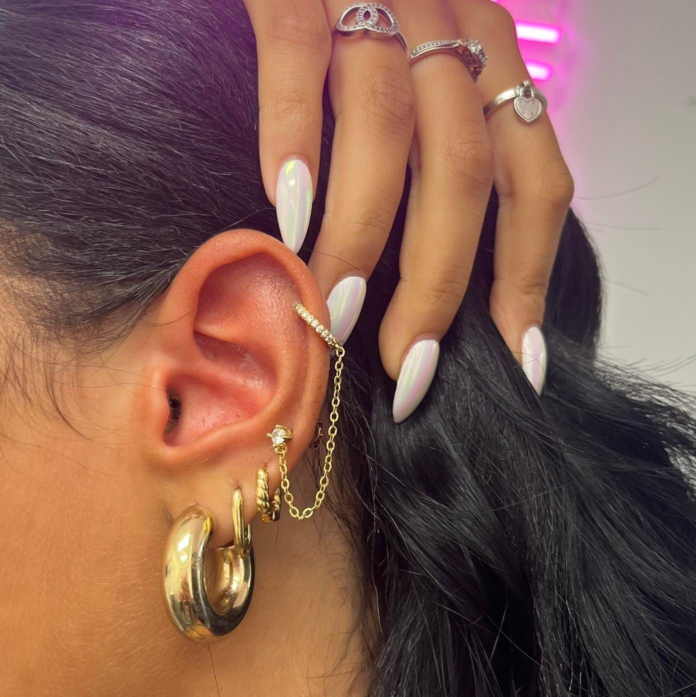 Woman's ear with gold jewelry, including hoops, ear cuff, and linked earrings. White nails, rings.