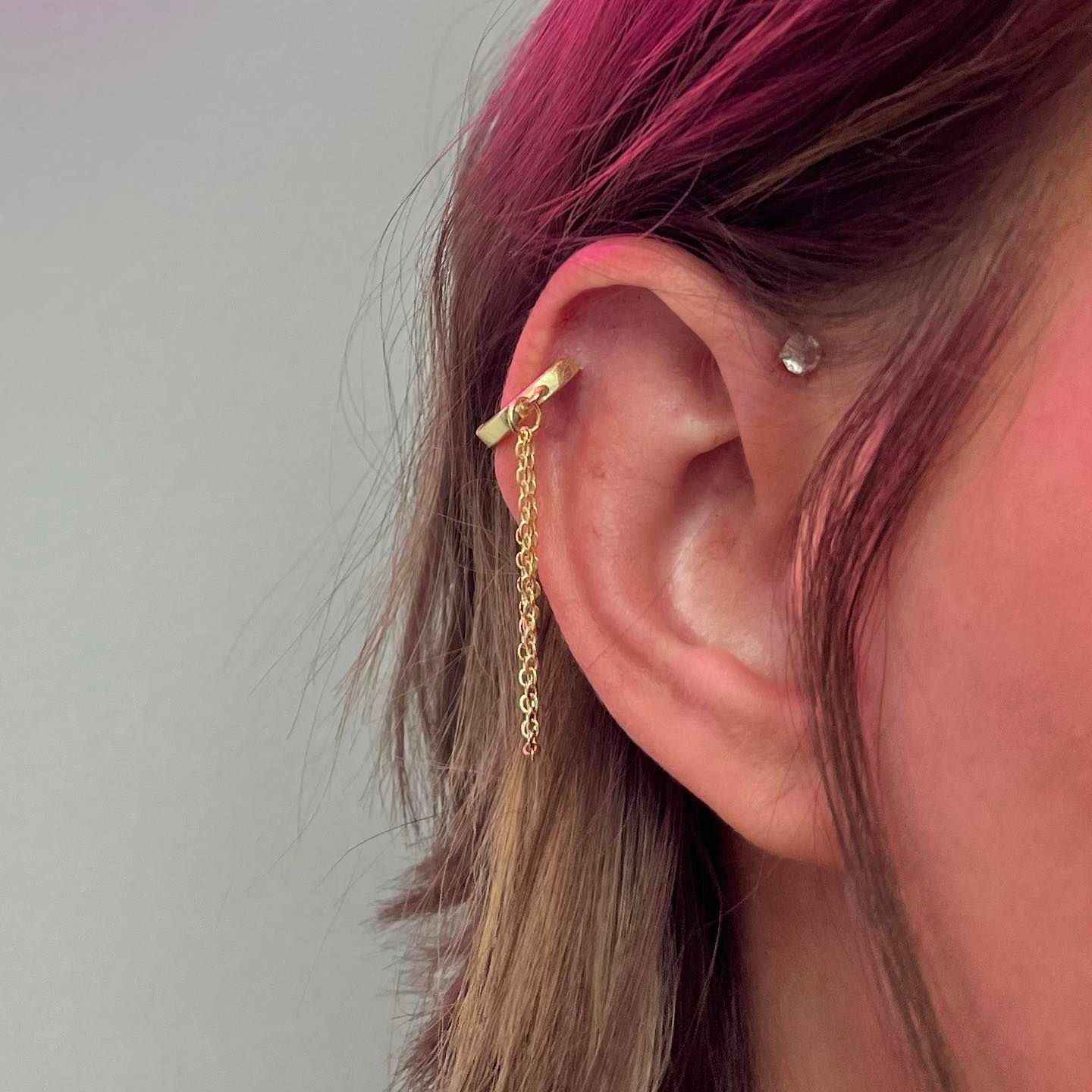 Ear with a gold chain earring and a small stud earring, pink highlights in hair.