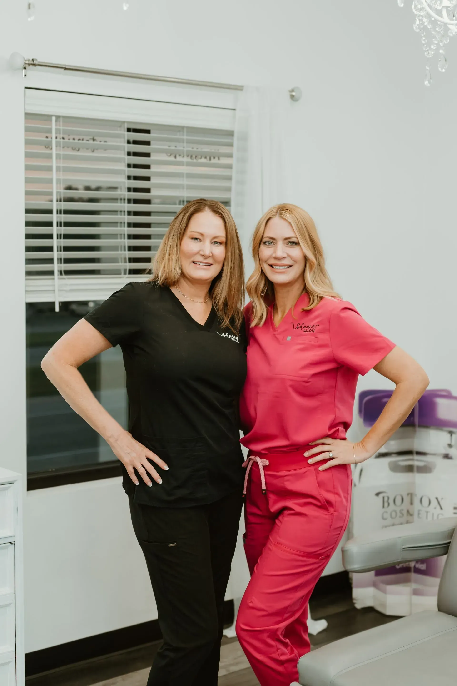 Two women in medical scrubs smiling, posing indoors. One in black, one in pink.