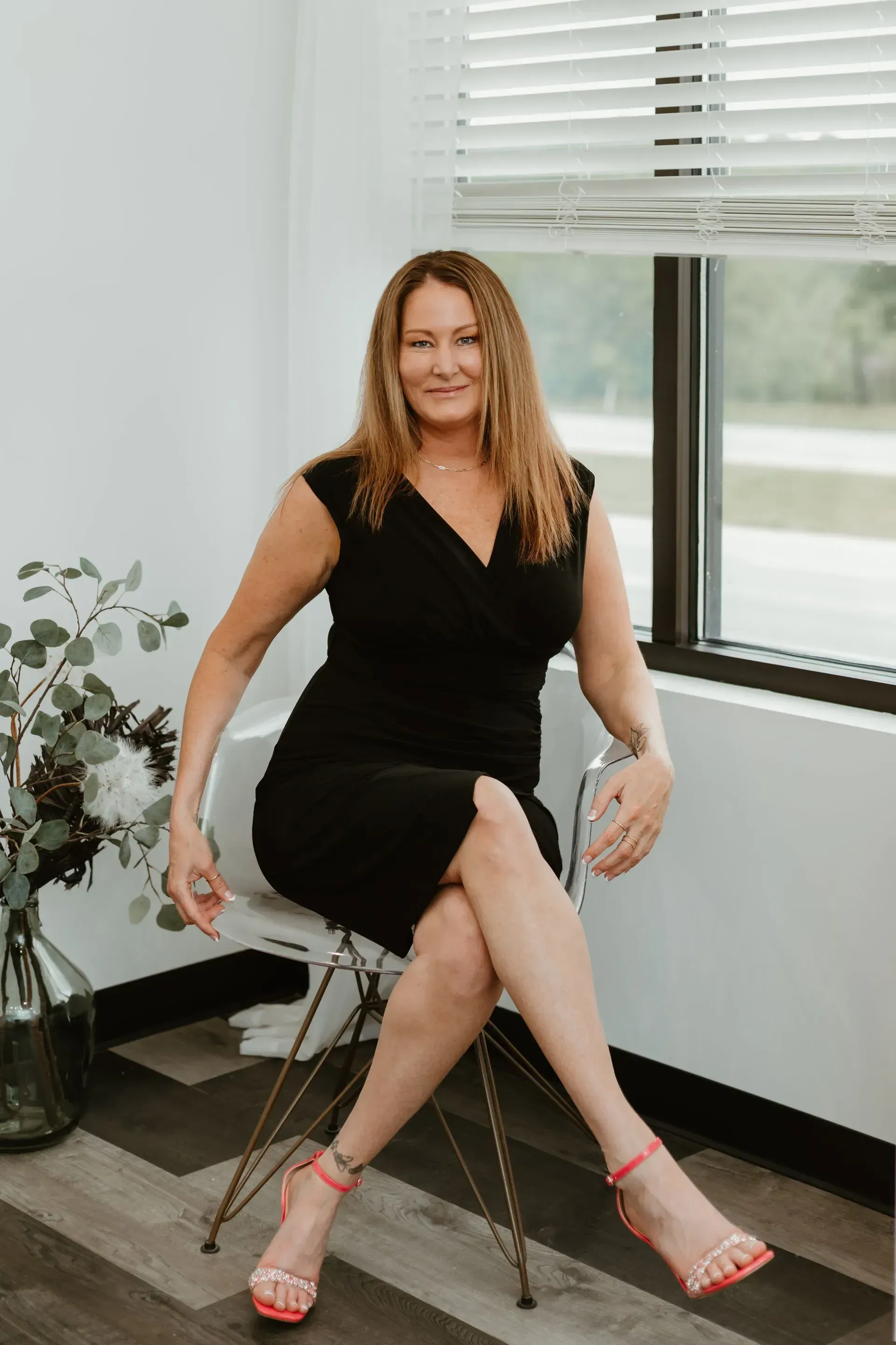 Woman in black dress sits on chair, legs crossed, pink heels, indoors, by a window.