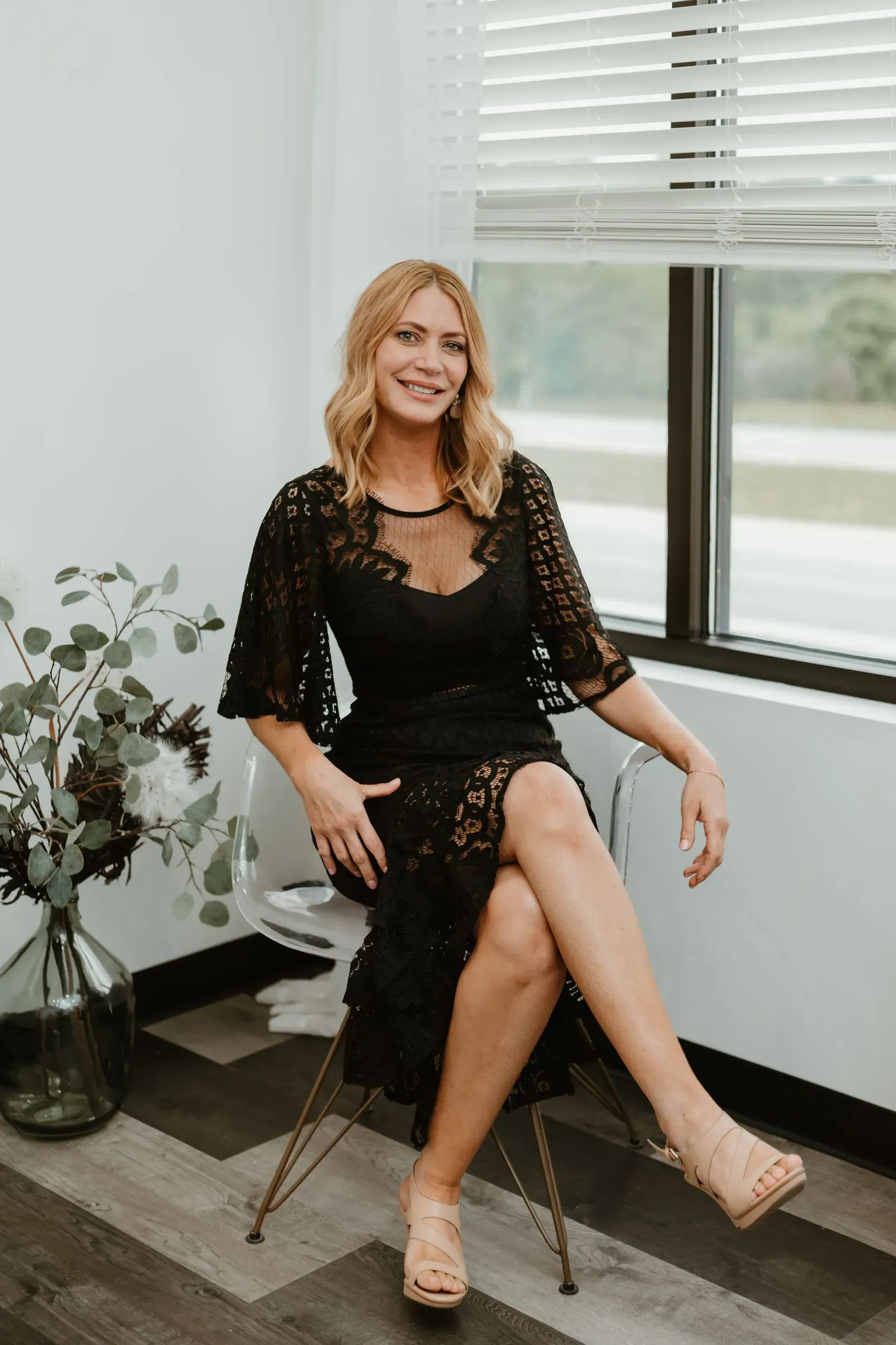 Woman in black lace dress sitting near a window, smiling.