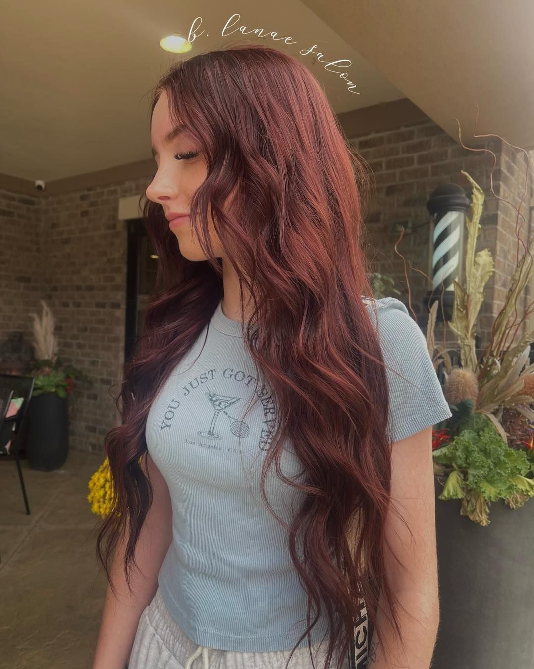 Woman with long, wavy, red hair. She wears a light blue shirt. Outdoors, near a barber shop.