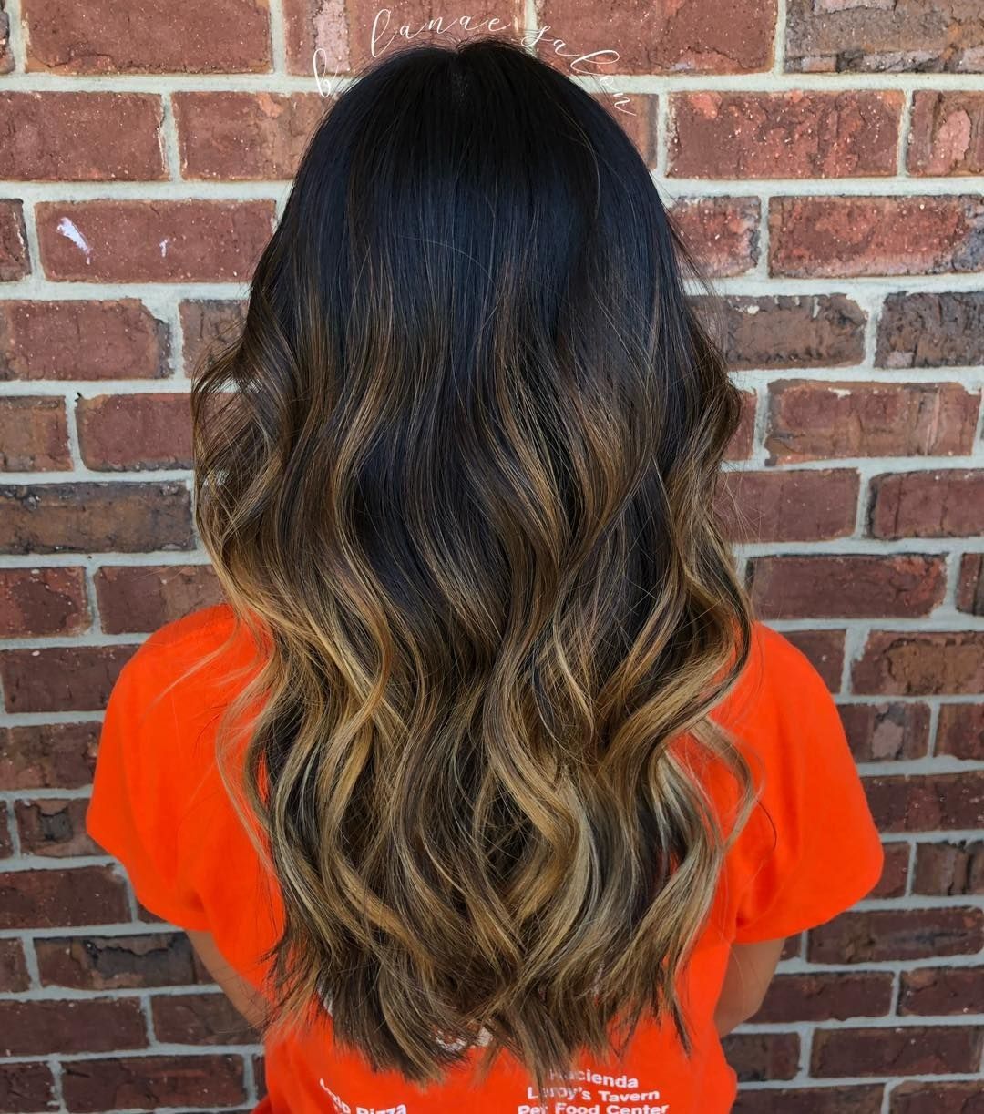 Woman with long dark hair, ombre style, against a brick wall, wearing an orange shirt.