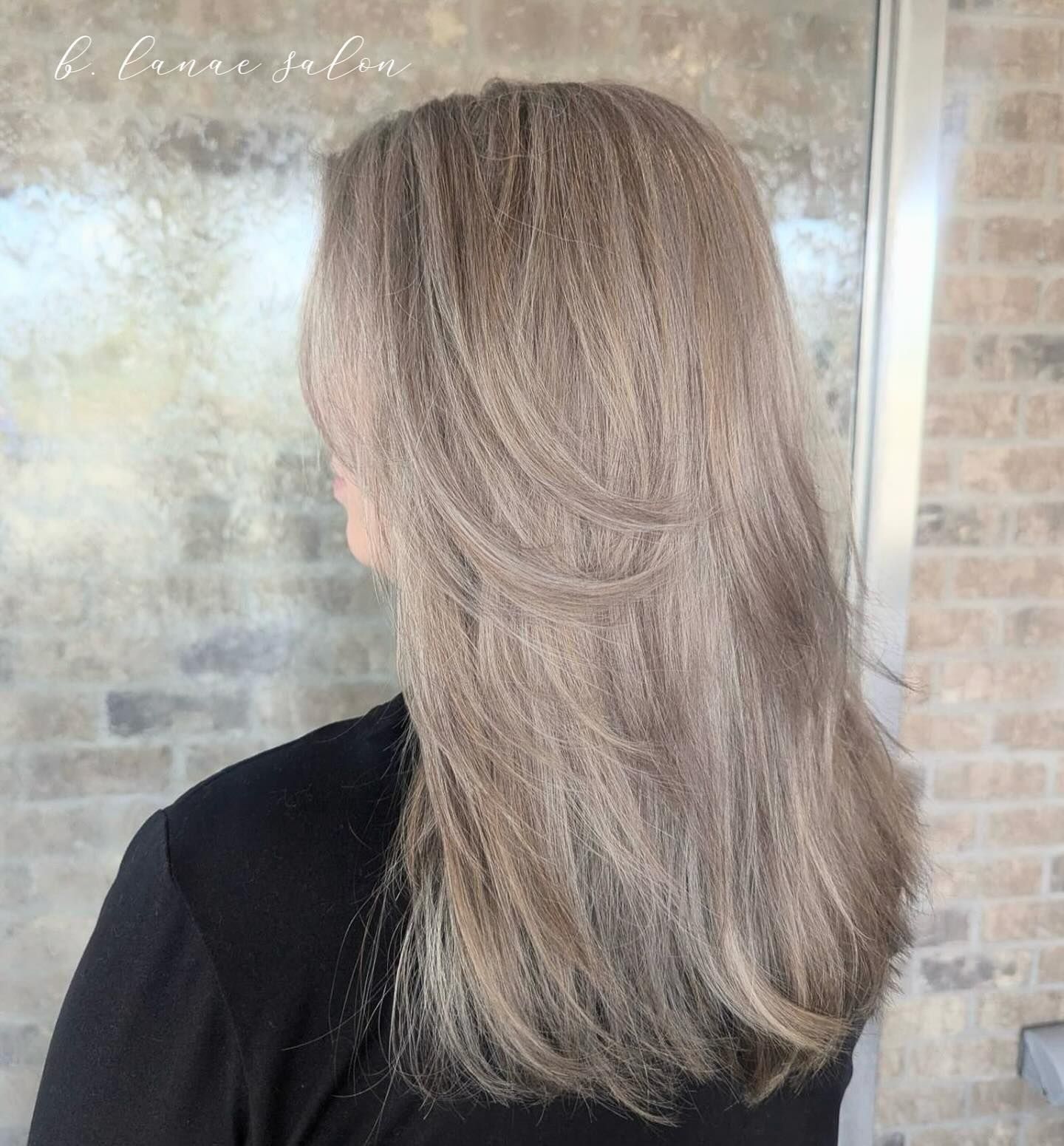 Woman with long, layered, ash-blonde hair, facing away from the camera. Outdoors, against a brick wall.