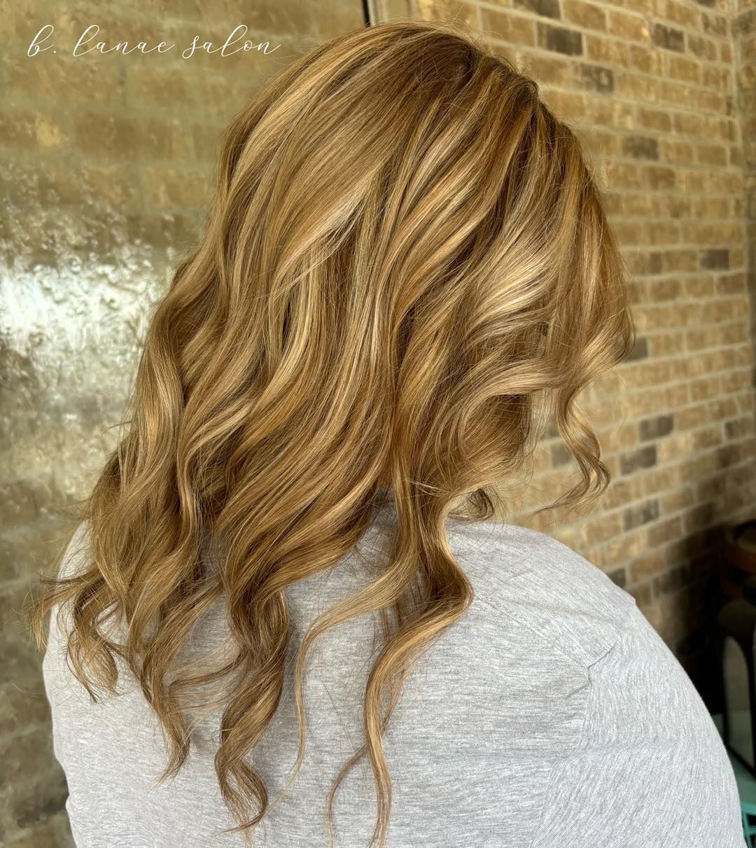 Woman with wavy, light brown hair, highlighted with lighter blonde, against a brick wall.