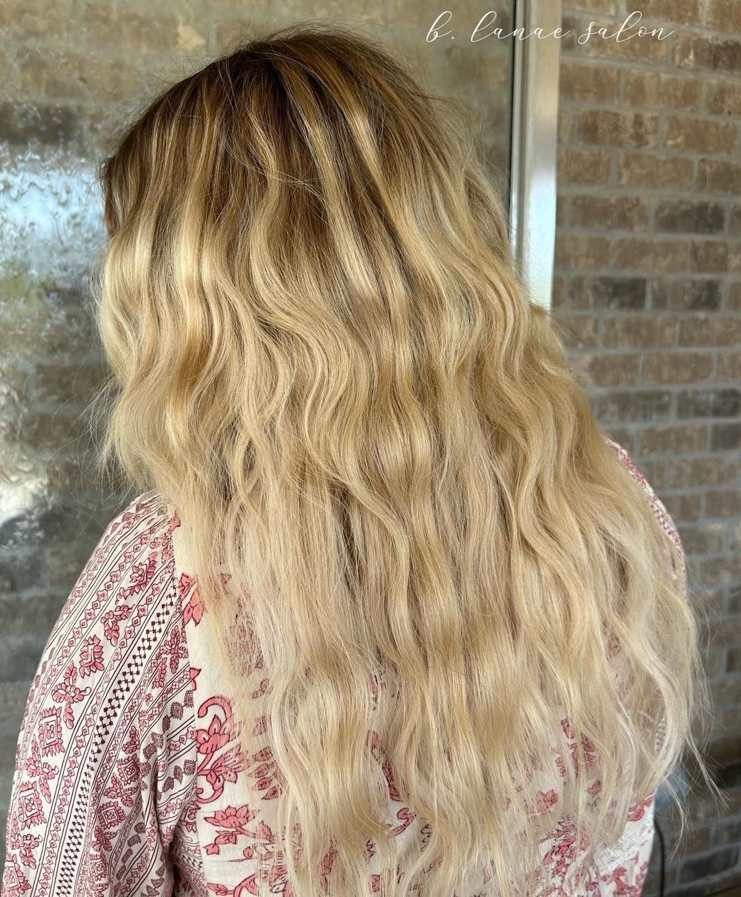 Woman with long wavy blonde hair, back view, wearing a patterned top.