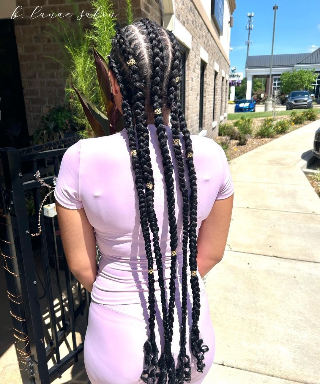 Woman with long braids, in a pink outfit, outdoors near a building and sidewalk.