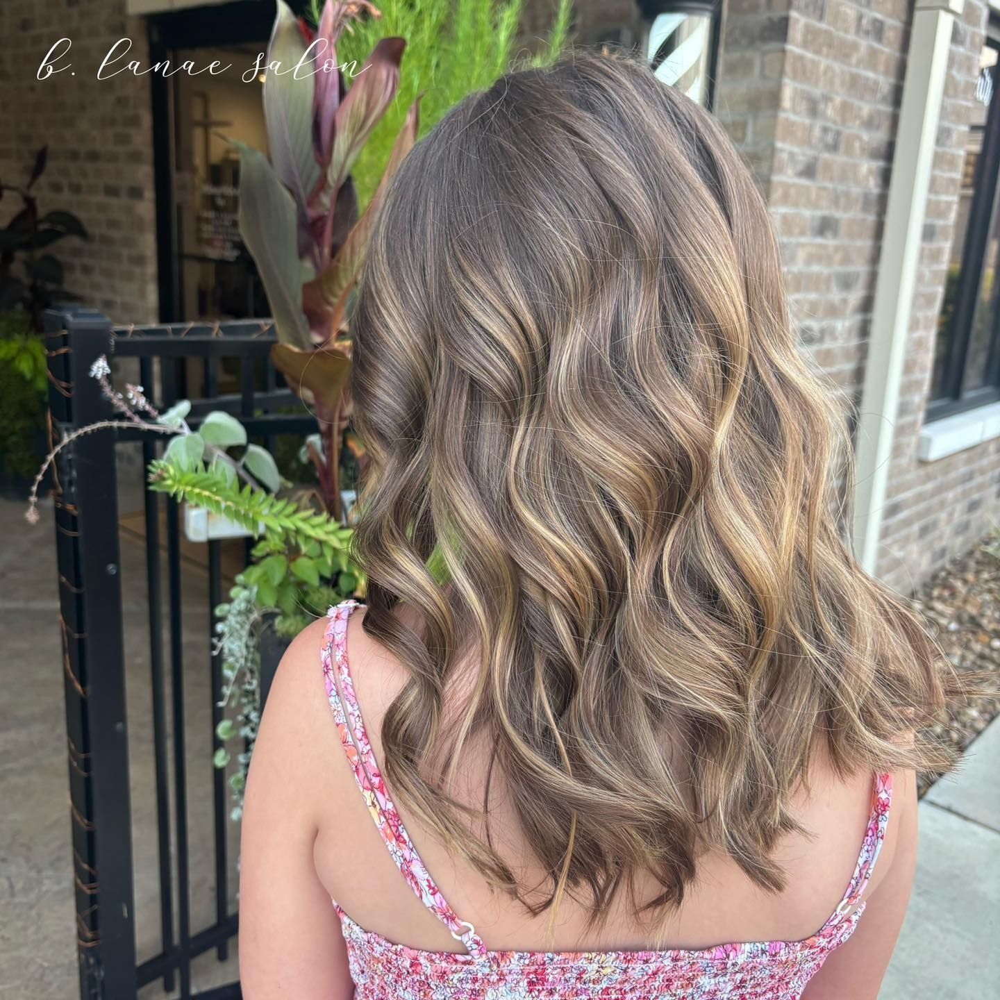Woman with wavy brown hair, highlighted with blonde streaks, outdoors near a brick building.