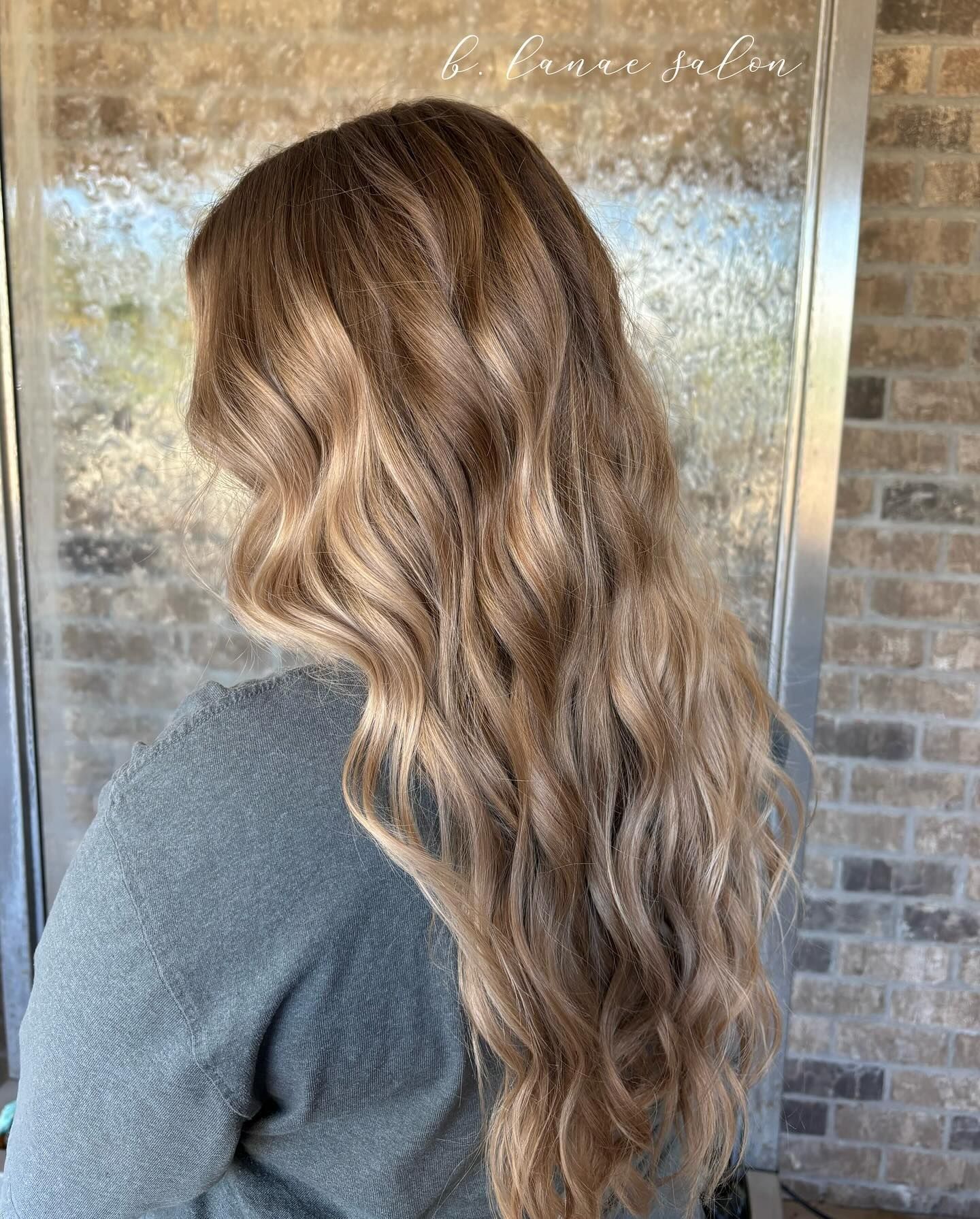 Woman with long wavy blonde hair, wearing a gray sweater, standing by a brick wall.