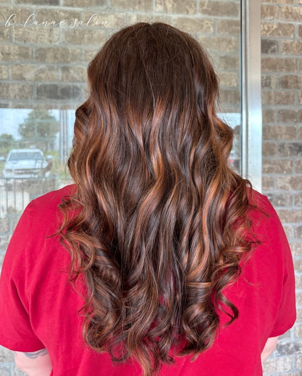 Woman with long, wavy auburn hair and red shirt, outdoors against a brick wall and window.