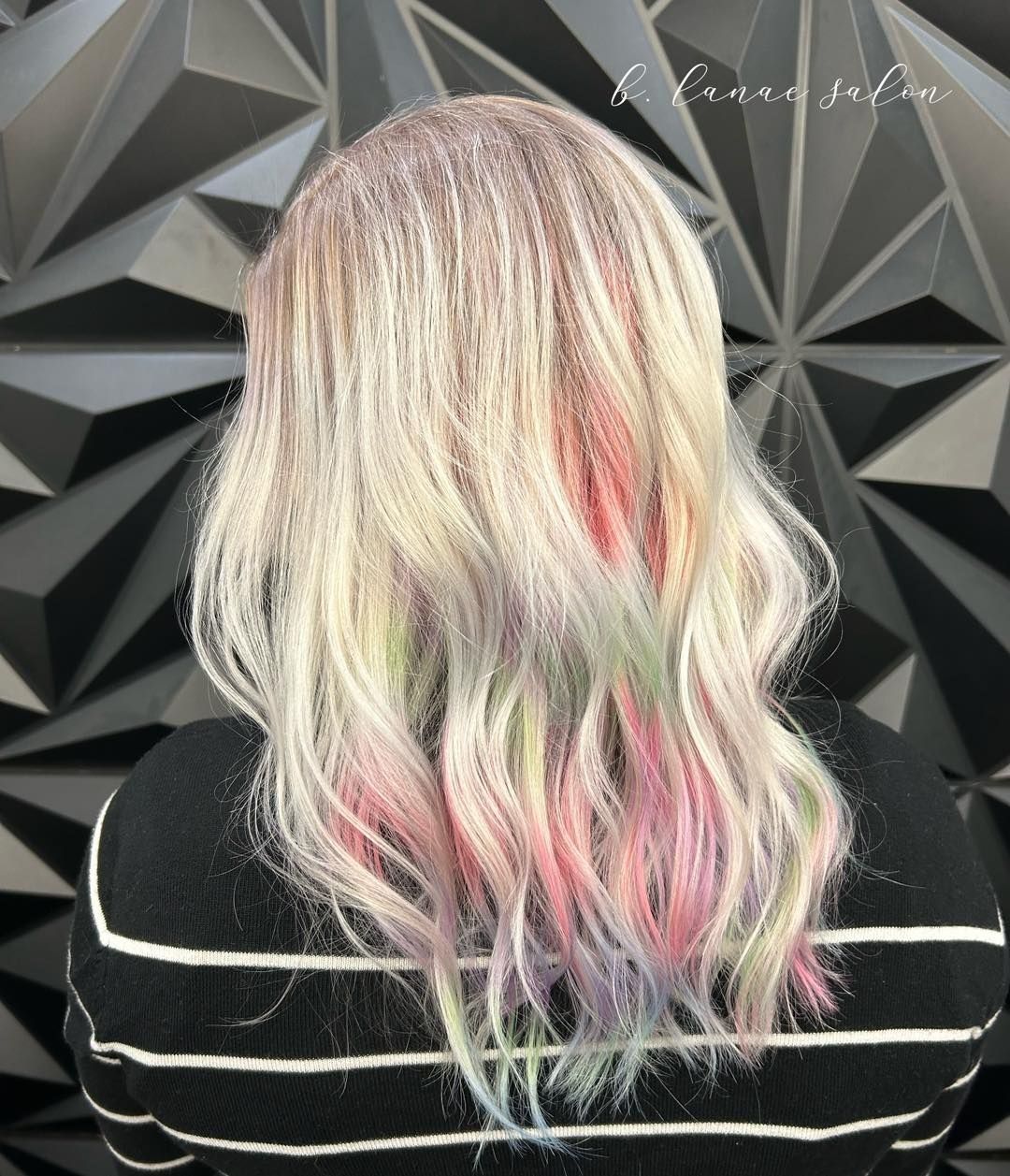 Woman with wavy, light blonde hair with rainbow streaks, standing in front of a geometric black wall.
