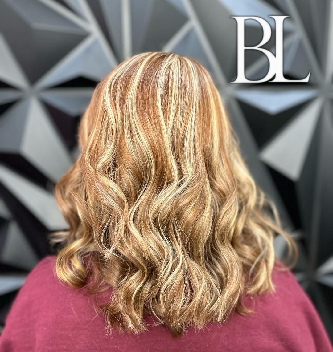 Woman with wavy auburn hair highlighted with blonde; background is a patterned gray wall.