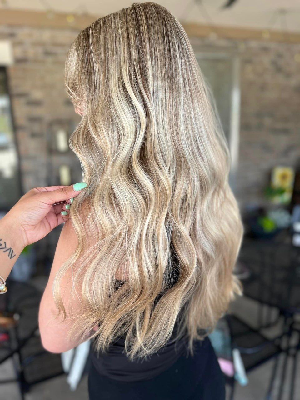 Woman with long blonde crimped hair, wearing a black top. Someone's hand touches her hair. Outdoor setting.