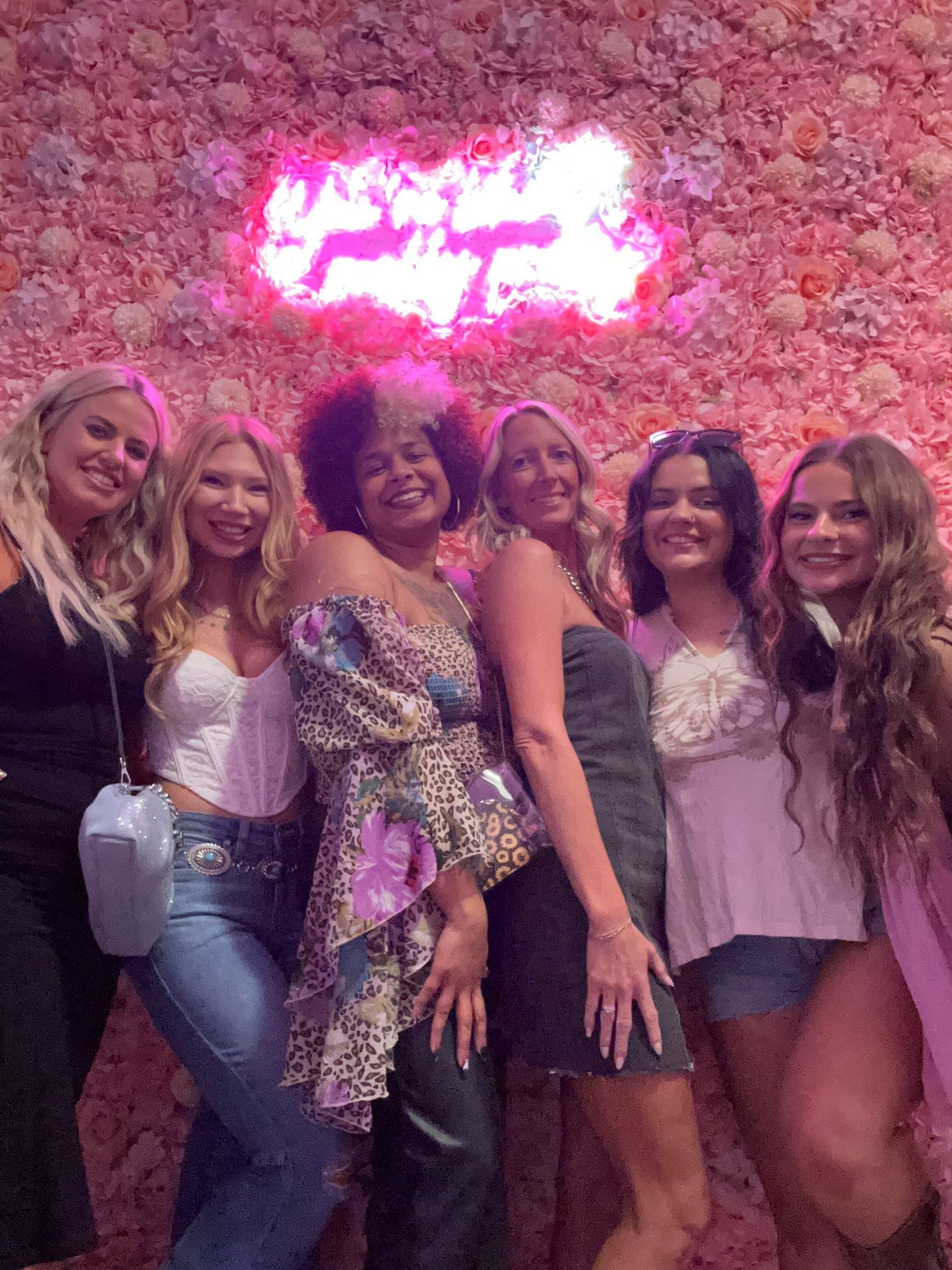 Six smiling women pose in front of a pink floral wall with a neon sign.